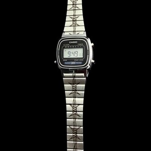 May include: A silver Casio digital watch with a black face displaying the time 9:49. The watch band features a repeating barbed wire design. The watch has the word "Casio" printed on the face.