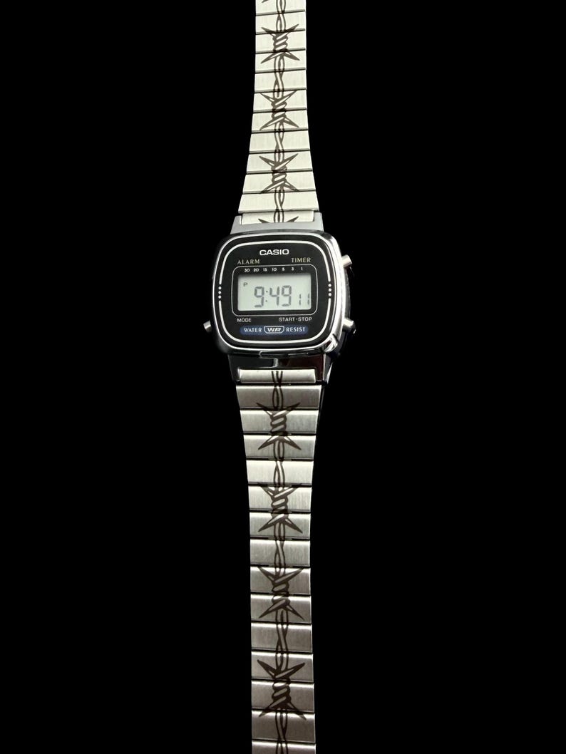 May include: A silver Casio digital watch with a black face displaying the time 9:49. The watch band features a repeating barbed wire design. The watch has the word "Casio" printed on the face.
