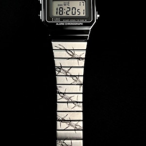 May include: A black Casio digital watch with a silver band featuring a barbed wire design. The watch face displays the time, day, and date. The watch has a water-resistant feature and alarm chronograph.