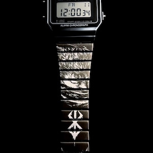 May include: Black Casio digital watch displaying the time 12:00:34. The watch band features a series of black and white graphic designs. The watch face includes the Casio logo and text: WATER RESIST, ALARM, SPL, CHRONO, LIGHT, MODE, START-STOP/12-24H, and ALARM CHRONOGRAPH.