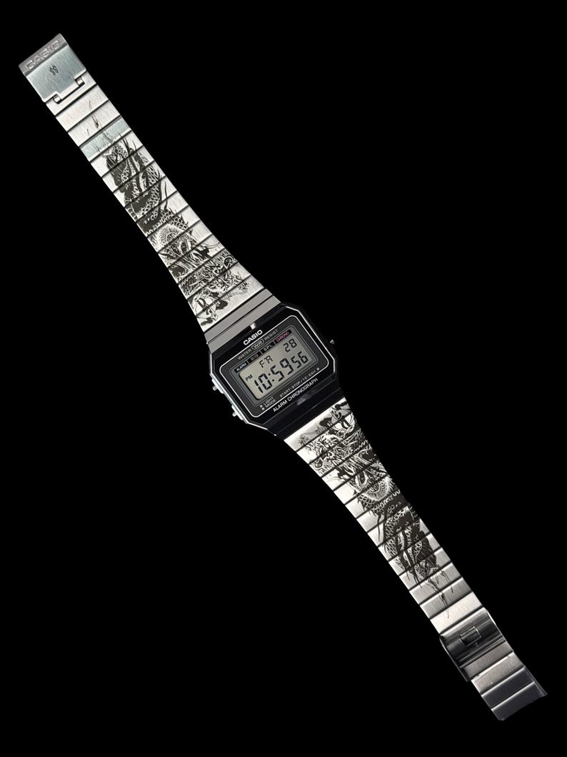Puede incluir: Un reloj de pulsera digital plateado con una esfera rectangular negra que muestra el logotipo de Casio, el texto Alarm Chronograph y una pantalla digital con la hora 10:59:56. La correa de eslabones met&aacute;licos tiene un dibujo de drag&oacute;n negro.