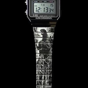 Può includere: Orologio digitale Casio nero con cinturino a tema Godzilla. Il quadrante mostra l'ora, il giorno e la data. Il cinturino presenta una grafica in bianco e nero di Godzilla. L'orologio ha il testo "CASIO" e "WATER WR RESIST".
