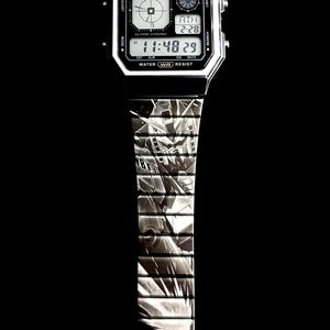 May include: A digital Casio watch with a black and silver design. The watch face displays the time and date. The watch band features a detailed graphic pattern. The watch has the text "CASIO ILLUMINATOR" and "WATER WR RESIST".