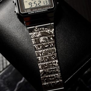 May include: A black digital Casio watch with a rectangular face and a metal link strap. The watch face displays the text CASIO, WATER RESIST, ALARM CHRONOGRAPH, and the time 9:52 13. The metal strap features an intricate, dark engraved pattern.