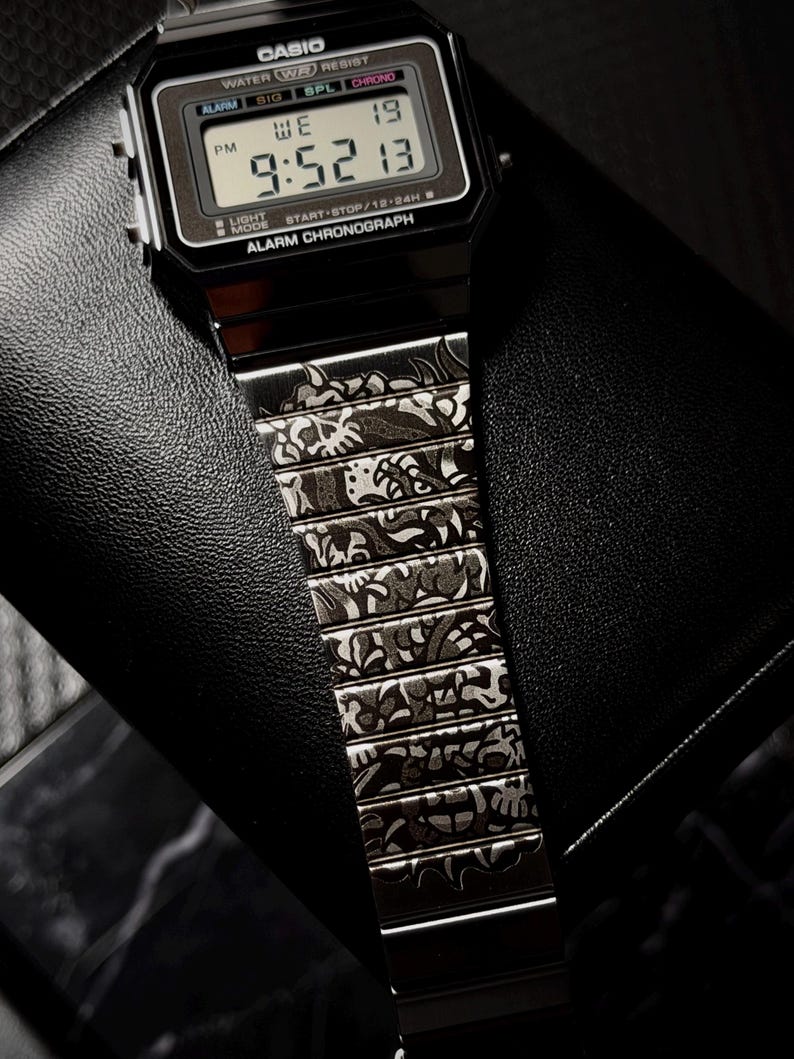 May include: A black digital Casio watch with a rectangular face and a metal link strap. The watch face displays the text CASIO, WATER RESIST, ALARM CHRONOGRAPH, and the time 9:52 13. The metal strap features an intricate, dark engraved pattern.