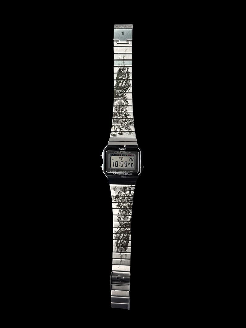 Puede incluir: Un reloj de pulsera digital plateado con esfera negra rectangular y correa de eslabones met&aacute;licos. La correa presenta una ilustraci&oacute;n detallada de un drag&oacute;n en estilo de tinta negra sobre los eslabones. La pantalla muestra 10:59:56 y FR 28.