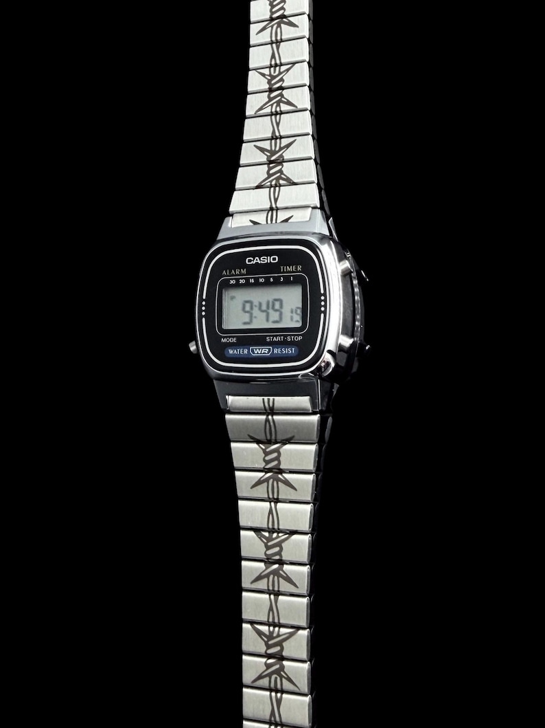 May include: A silver Casio digital watch with a black face displaying the time 9:49. The watch band features a barbed wire design. The watch has alarm, timer, mode, start-stop, water resist functions.
