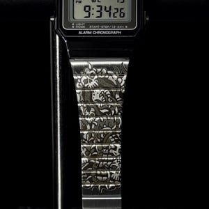 May include: A black digital wristwatch with a rectangular face featuring the text CASIO, WATER RESIST, ALARM, and CHRONOGRAPH. The digital display shows the time 9:34 and the date 19. The metal watch band is engraved with a detailed skull pattern.