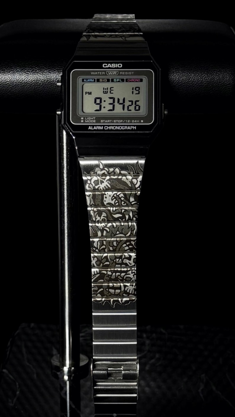 May include: A black digital wristwatch with a rectangular face featuring the text CASIO, WATER RESIST, ALARM, and CHRONOGRAPH. The digital display shows the time 9:34 and the date 19. The metal watch band is engraved with a detailed skull pattern.