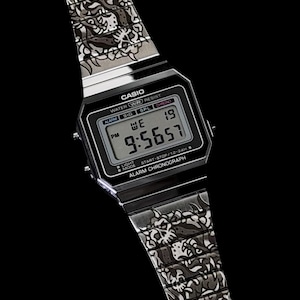 May include: A silver-tone digital wristwatch featuring a rectangular black face with the text CASIO, WATER WR RESIST, and ALARM CHRONOGRAPH. The metal band displays a detailed, repeating dark graphic pattern of stylized skulls and organic shapes.