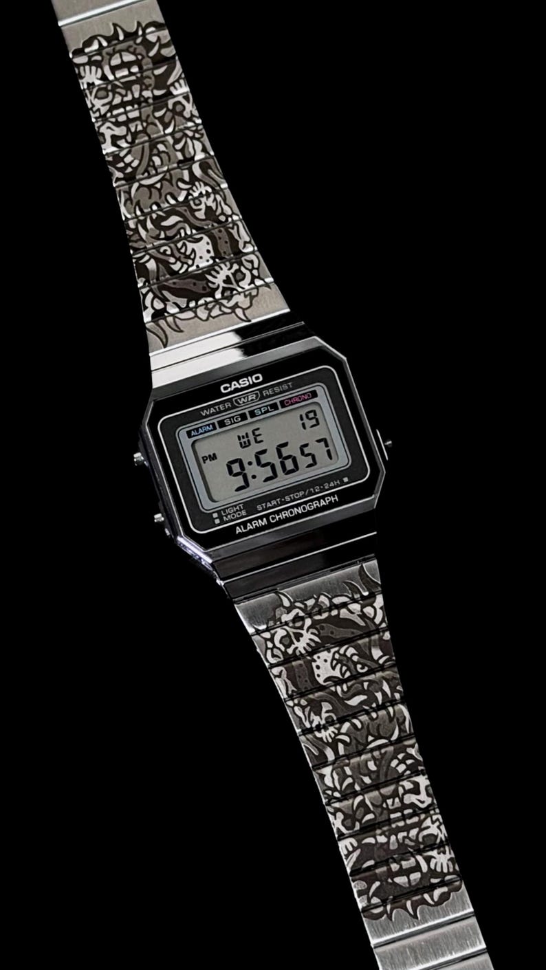 May include: A silver-tone digital wristwatch featuring a rectangular black face with the text CASIO, WATER WR RESIST, and ALARM CHRONOGRAPH. The metal band displays a detailed, repeating dark graphic pattern of stylized skulls and organic shapes.