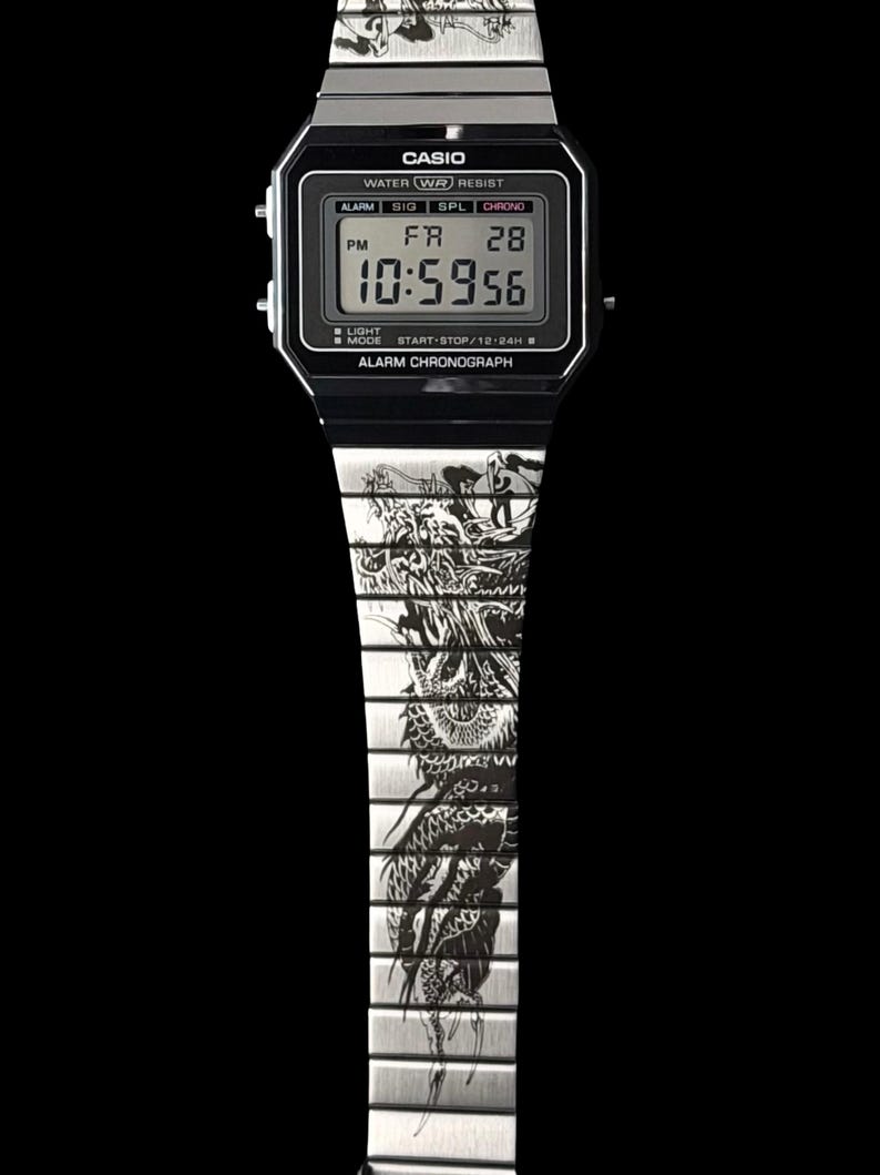 Puede incluir: Un reloj de pulsera digital plateado con una esfera rectangular negra que muestra el logotipo de Casio y el texto alarm y chronograph. La correa de eslabones met&aacute;licos presenta un grabado detallado de un drag&oacute;n negro en los eslabones.