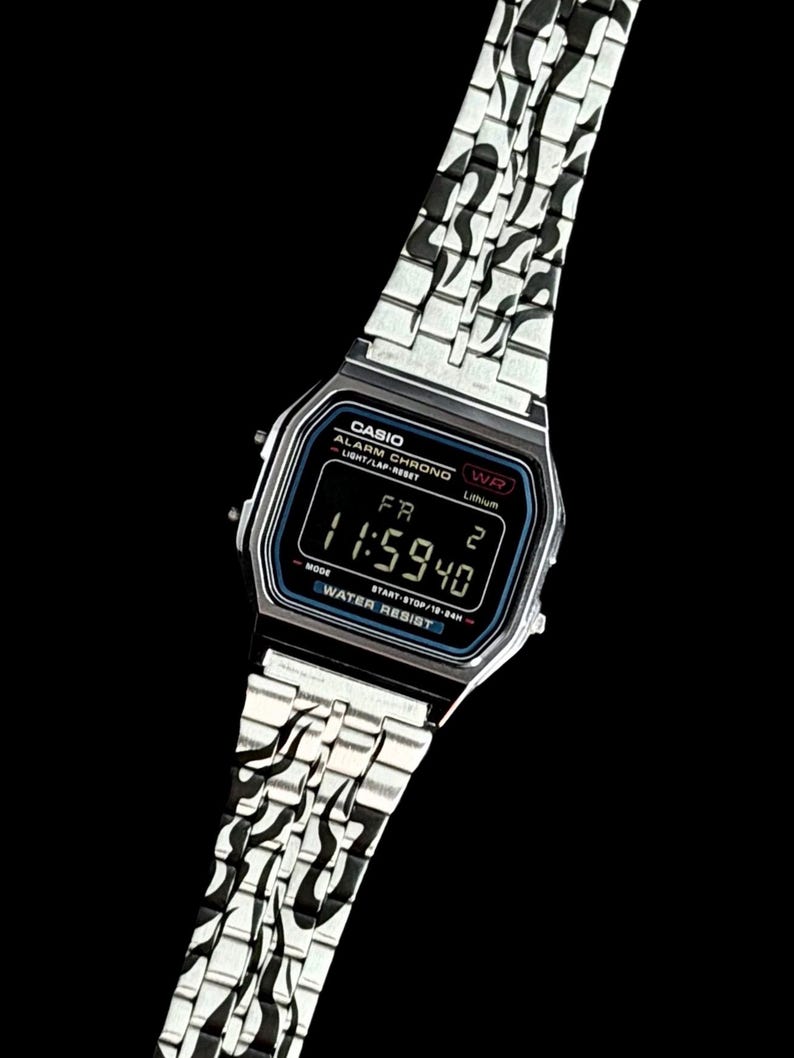 May include: A silver-tone digital wristwatch with a black face and a metal link band featuring a black flame pattern. The watch display shows the text CASIO, ALARM CHRONO, WR, Lithium, WATER RESIST, and the time 11:59:40 with the day FR 2.