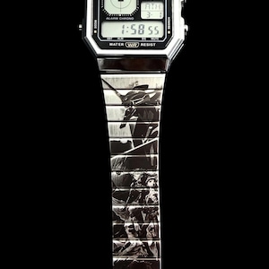 Puede incluir: Un reloj digital Casio plateado con una esfera negra que muestra la hora 1:58:55. La correa del reloj presenta un diseño gráfico en blanco y negro con la palabra "EVANGELION" al final. El reloj tiene el texto "CASIO ILLUMINATOR" en la parte superior.