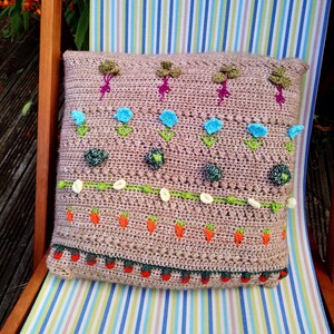 May include: A brown crocheted pillow with a floral design. The pillow features green leaves, blue flowers, and orange carrots. The pillow is decorated with a white and green border.