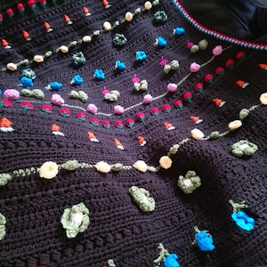 May include: A black crocheted blanket with a floral design. The blanket features a variety of colorful flowers, including red, blue, pink, and yellow. The blanket is made with a textured stitch that gives it a unique look.