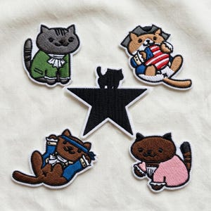 May include: Embroidered patches of cartoon cats in historical attire. The patches depict a grey cat in a green coat, a brown cat with an American flag, a brown cat with a blue headband, and a brown cat in a pink outfit. A black star is in the centre.