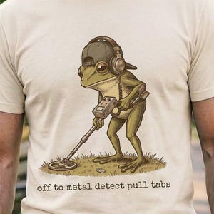 Puede incluir: Camiseta color marfil con una rana de dibujos animados con gorra y auriculares, usando un detector de metales. El texto "off to metal detect pull tabs" está impreso debajo. La camiseta es de material suave.