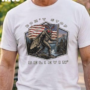 May include: A white t-shirt featuring a graphic of a moth-like creature holding an American flag. The design is enclosed in a hexagon shape with a landscape background and the text "DON'T STOP BELIEVIN'" printed in a dark font above and below the image.