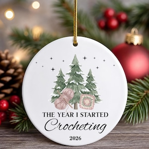 May include: A round white ceramic ornament with a gold hanging cord. The design features three green pine trees, a ball of pink yarn, and a square crochet motif. The text reads: THE YEAR I STARTED Crocheting 2026 in black lettering.