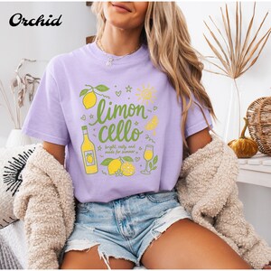 May include: A light purple short-sleeve t-shirt featuring a graphic print of lemons, a bottle, and a glass with the text "limon cello" and "bright, zesty, and made for summer" in green script, accompanied by small sun, star, and heart icons.