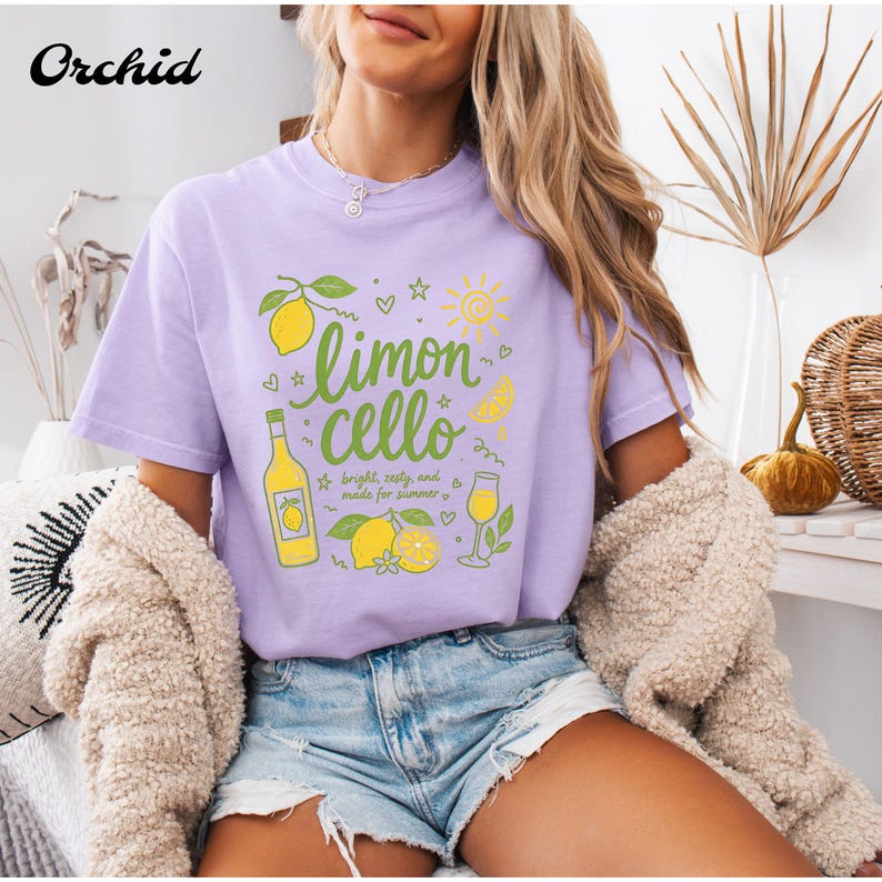 May include: A light purple short-sleeve t-shirt featuring a graphic print of lemons, a bottle, and a glass with the text "limon cello" and "bright, zesty, and made for summer" in green script, accompanied by small sun, star, and heart icons.