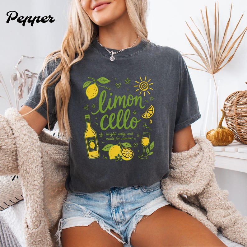 May include: A charcoal gray short-sleeve t-shirt featuring a yellow and green graphic print of lemons, a bottle, and a glass with the text "limoncello" and "bright, zesty, and made for summer" written in a cursive and block font style.