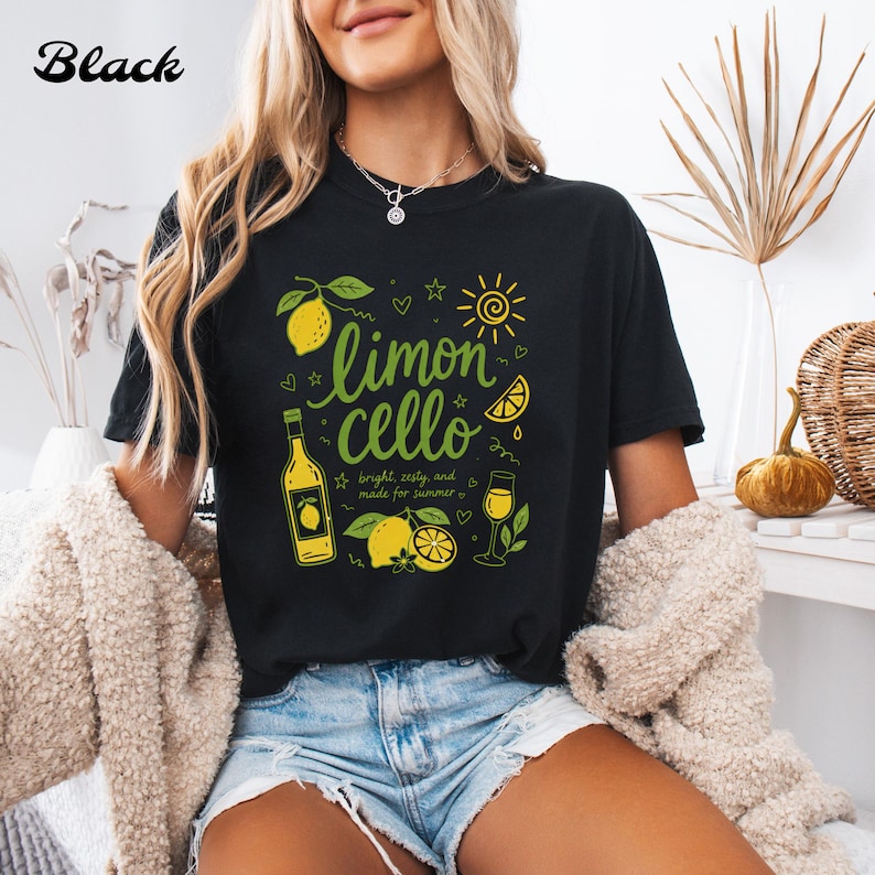 May include: A black t-shirt featuring a graphic print with the text "limoncello" in green, accompanied by illustrations of lemons, a bottle, a glass, and a sun. The design includes the text "bright, zesty, and made for summer" in small font.