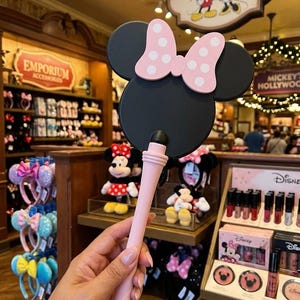 May include: A handheld mirror featuring a black circular frame shaped like a mouse head, topped with a light pink bow decorated with white polka dots. The handle is a solid light pink, tapering slightly toward the base with a smooth, matte finish.
