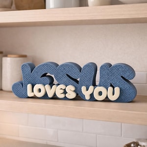 May include: A blue decorative sign with the words "JESUS LOVES YOU" in a stylized font. The word "JESUS" is in a textured blue, while "LOVES YOU" is in a cream color. The sign is displayed on a wooden shelf.