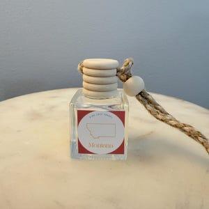May include: A small, clear glass bottle with a wooden cap and a rope loop. The bottle has a red and white label with the word "Montana" and a state outline. The rope is beige and white. The text "KIDS LOVE TRAVEL" is on the label.