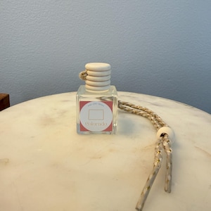 May include: A clear glass car air freshener with a wooden cap and a hanging rope. The label displays a pink rectangle with a white outline of Colorado and the state name. The rope is beige with gold detailing.