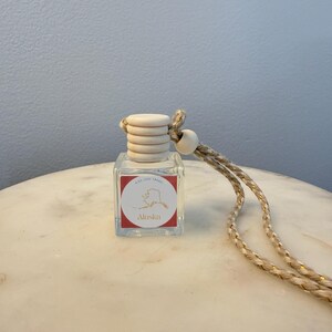 Op de afbeelding: Een kleine, heldere glazen fles met een houten dop en een decoratief touw. De fles heeft een rood etiket met het woord "Alaska" en een kaartomtrek. Het touw is beige met gouden accenten. De fles is ontworpen om op te hangen.