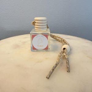 May include: A small, clear glass bottle with a wooden cap and a decorative cord. The bottle has a label with the outline of Maine and the word "Maine". The cord is beige with gold detailing.