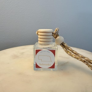 Puede incluir: Una pequeña botella de vidrio transparente con una tapa de madera y un cordón decorativo. La botella tiene una etiqueta roja y blanca con la palabra "Nebraska" y un contorno del estado. El texto "Ride Love Travel" también está presente.