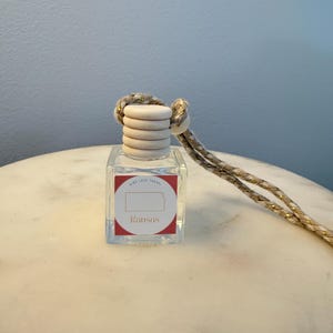 Könnte beinhalten: Eine klare, quadratische Glas-Lufterfrischerflasche mit einem Holzdeckel und einem dekorativen Seil. Das Etikett zeigt einen roten Kreis mit dem Bundesstaat Kansas und dem Wort "Kansas". Das Seil ist beige mit goldenen Akzenten.