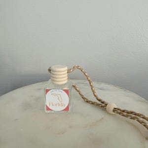 May include: A small, clear glass bottle with a wooden cap and a Florida-shaped graphic on a red square label. The bottle hangs from a tan and gold rope. The product is sitting on a marble surface.