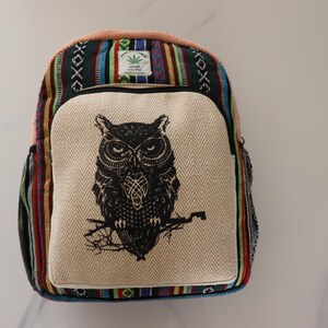 May include: A colourful backpack with a large owl graphic on the front. The bag features a beige front panel with a black owl illustration, and a striped pattern in various colours. The top has a hemp leaf patch.