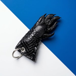 May include: A black, claw-shaped keychain with a textured, reptilian-like pattern. The keychain features a silver metal ring for attaching keys. The background is divided diagonally, with a white and blue color scheme.