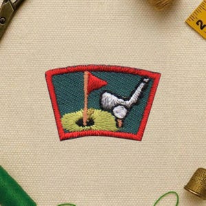 May include: Embroidered patch featuring a golf scene. The patch has a green background with a red border. It depicts a golf ball, club, flag, and hole. The flag is red, and the golf ball is white.