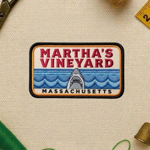 May include: Embroidered patch featuring the text "MARTHA'S VINEYARD" in red, above a blue ocean scene with a shark and the text "MASSACHUSETTS". The patch has a black border with an orange outline.