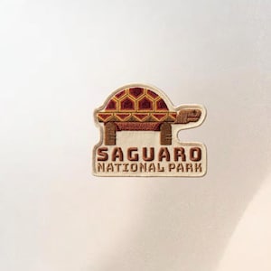 May include: Embroidered patch featuring a turtle design with a geometric pattern on its shell. The patch includes the text "SAGUARO NATIONAL PARK" in a brown, textured font, and is bordered by a beige outline.