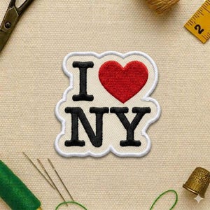 May include: Embroidered patch with the text "I ❤️ NY" in black letters and a red heart. The patch has a white border and is on a beige fabric background. Sewing tools, including scissors, needles, and thread, are visible.