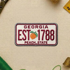 May include: Embroidered patch in the shape of a Georgia license plate. The patch features the words "GEORGIA" at the top, "EST 1788" in burgundy, and a peach graphic. The words "PEACH_STATE" are at the bottom.