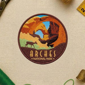 May include: Embroidered patch featuring a scenic view of Arches National Park. The patch has a brown border and depicts a landscape with arches, a deer, and the text "ARCHES NATIONAL PARK". The design incorporates shades of brown, orange, green, and blue.