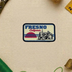 May include: Embroidered patch featuring the word "FRESNO" in blue, with "California" in red. A tractor graphic is below a red and blue sunset design. The patch has a blue border and is on a beige background.