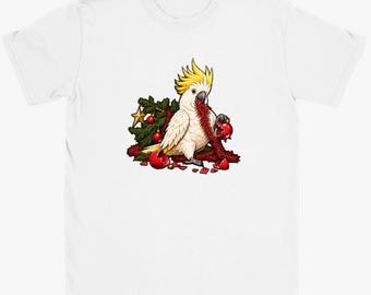May include: A white short-sleeved t-shirt featuring a graphic of a white cockatoo with a yellow crest. The bird is surrounded by green pine branches, red Christmas baubles, and red tinsel, with some baubles appearing broken.