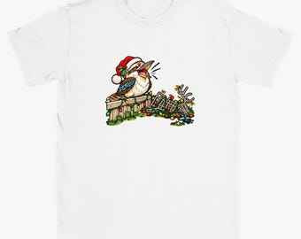 May include: A white short-sleeve t-shirt featuring a graphic of a kookaburra wearing a red and white Santa hat. The bird sits on a wooden fence draped with colourful Christmas lights, with a fallen reindeer antler decoration nearby.