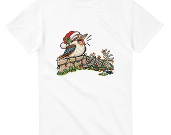 May include: A white short-sleeve t-shirt featuring a graphic of a kookaburra wearing a red and white Santa hat. The bird sits on a wooden fence draped with colourful Christmas lights, with a tangled string of lights and a small antler nearby.