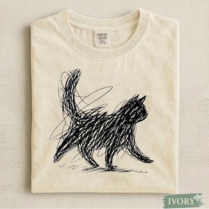 May include: Ivory-colored t-shirt featuring a black scribble-style cat illustration. The cat is depicted in a walking pose with its tail raised. The shirt has a crew neck and short sleeves. The tag reads "Comfort Colors".